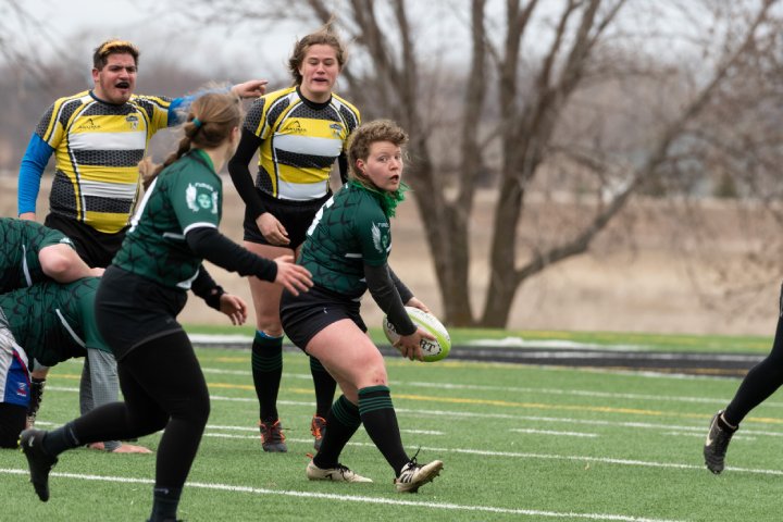 
              A rugby player in a green and black jersey holds the ball while looking for an opportunity to pass during a game. Teammates and opponents are positioned around, with a player in a yellow and black jersey pointing in the background. The action takes place on a turf field.
